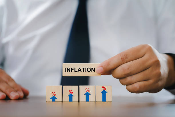 A person in formal attire holding a wooden block labeled inflation above three wooden blocks with upward arrows and percentage symbols, representing rising inflation rates.