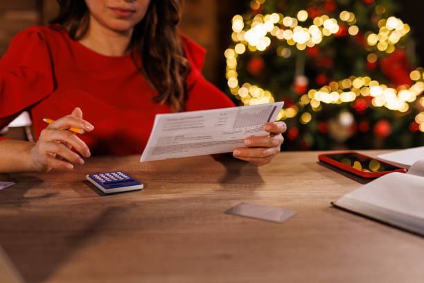 A mid-adult woman sits at her home office desk, stressed as she sorts through receipts, bills, and finances, concerned about managing holiday debt during the Christmas season.