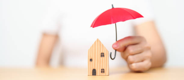 Person holding a small umbrella shielding a house, symbolizing protection and the importance of insurance against natural disasters and rising risks.